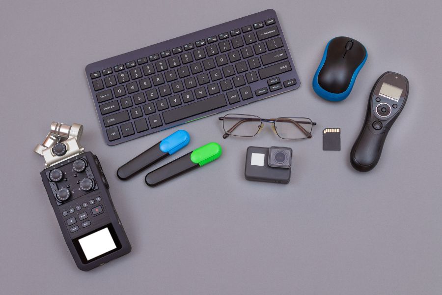 Une collection de gadgets high-tech disposés sur fond gris incluant clavier sans fil, souris, GoPro, enregistreur audio, télécommande et carte mémoire.